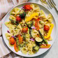 A close-up of vibrant Rainbow Veggie Pasta Primavera in a white bowl, showcasing colorful bow-tie pasta tossed with fresh spring vegetables and herbs.